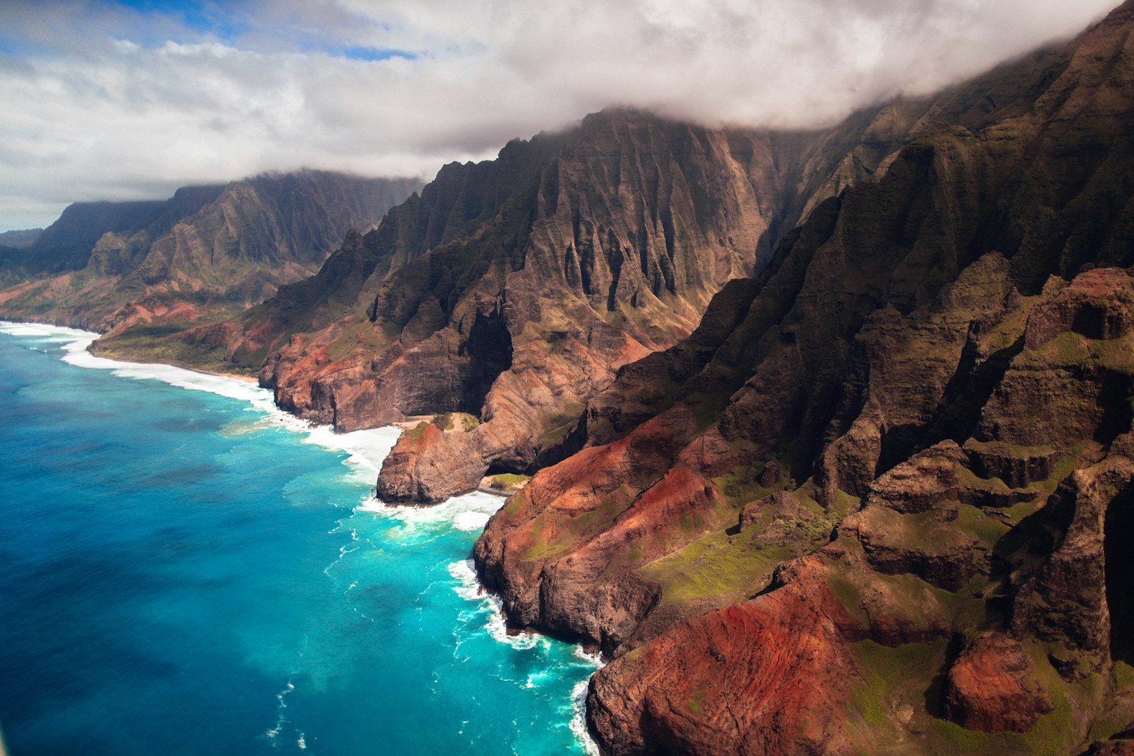 Aerial view of ocean waves breaking against steep green sea cliffs along a remote coastline.